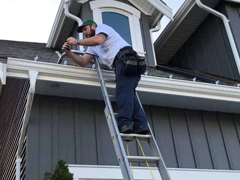 Installer on a ladder clipping C9 LED lights along a roofline
