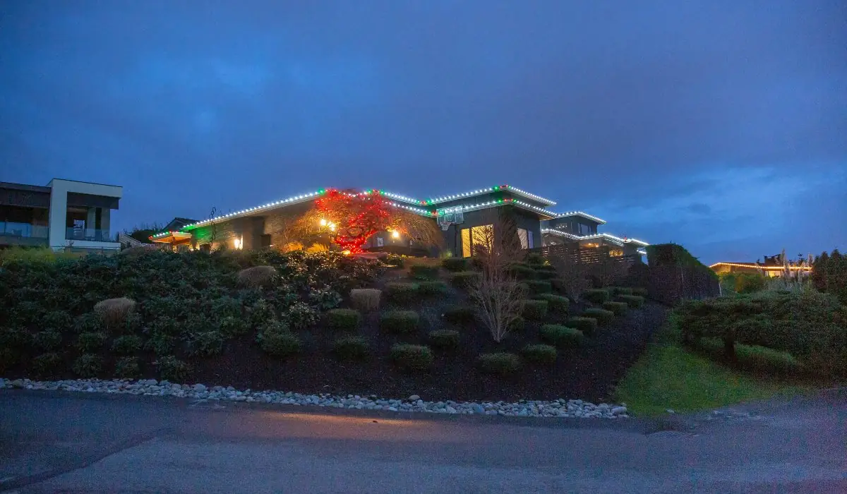 Low modern home with warm white roofline lights and red-lit shrubs in Yarrow Point, WA